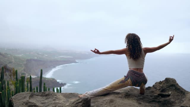 A-woman-sitting-on-the-edge-of-a-cliff-in-a-pose-war-overlooking-the-ocean-raise-her-hands-up-and-inhale-the-sea-air-while-doing-yoga