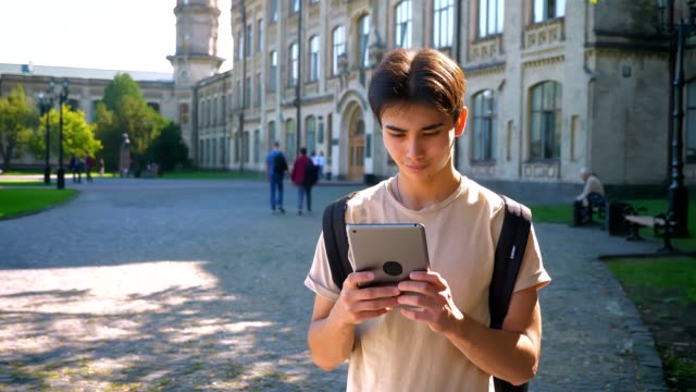 Concentrated-nice-asian-boy-is-using-his-tablet-while-standing-in-old-amazing-city,-spots-of-sunlight-in,-summer-mood