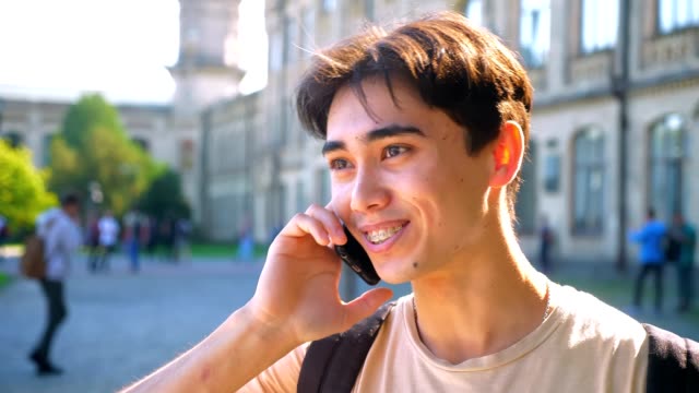 Nice-asian-boy-is-on-the-phone,-smiling-and-talking,-bright-sun,-urban-view-as-background