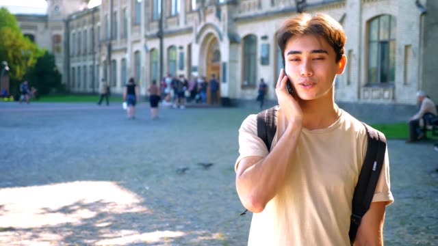 Pretty-asian-guy-is-talking-over-phone-and-looking-at-camera-straight-while-standing-near-park,-outdoor,-sunny-weather-in-city