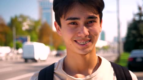 Shy-sincere-smile-of-asian-man-in-braces-looking-straight-at-camera,-showing-braces-while-standing-on-the-road-background,-urban-view