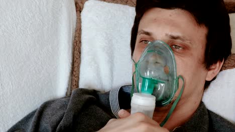 Use-nebulizer-and-inhaler-for-the-treatment.-Closeup-man's-face-inhaling-through-inhaler-mask-lying-on-the-couch.-Front-view