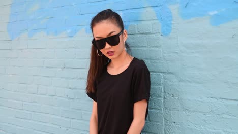 Young-beautiful-asian-girl-wearing-sunglasses-and-smiling-posing-near-wall-outdo