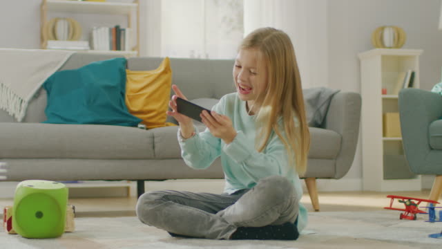 Chica-linda-inteligente-sentado-sobre-una-alfombra-jugando-en-juego-de-Video-en-su-teléfono-inteligente,-tiene-y-usa-teléfono-móvil-en-modo-apaisado-Horizontal.-Niño-tiene-diversión-jugando-videojuegos-en-la-sala-de-estar-soleado.