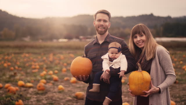 Portrait-of-a-young-family-at-a-pumpkin-patch,-mom-and-dad-holding-pumpkins,-with-lens-flare