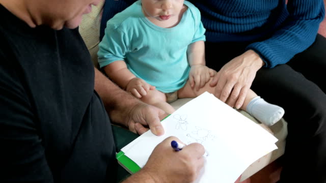 Attractive-baby-boy-draws-a-pen-with-his-grandparents-home-on-the-couch.-The-boy-stares-at-the-animals-that-drew-grandfather.-The-concept-of-different-generations