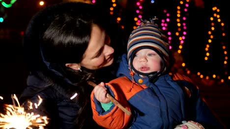 Young-mother-with-a-child-celebrate-the-new-year-on-the-street-with-a-sparkler.-Hugging-and-smiling-at-the-camera.-Against-the-background-is-decorated-Christmas-tree