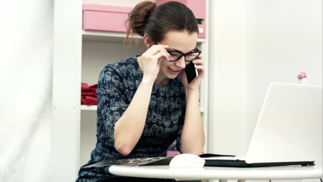 Young-attractive-female-fashion-designer-using-computer-laptop-and-talking-on-mobile-phone-call-in-manufacturing-office-studio