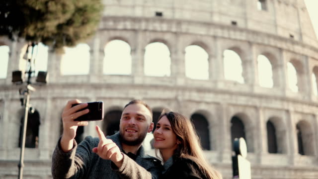 Feliz-pareja-joven-con-smartphone-para-tomar-fotos-selfie-cerca-de-Coliseo-de-Roma,-Italia.-Hombre-y-mujer-tienen-vacaciones