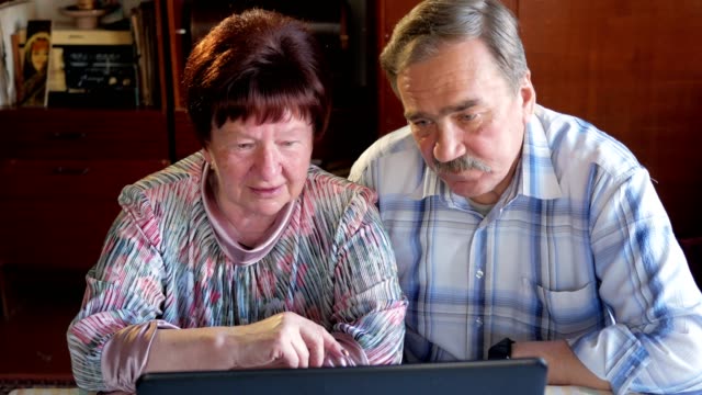 Una-pareja-de-ancianos-está-sentado-en-casa-en-la-computadora-portátil.-Una-mujer-lee-noticias,-un-hombre-con-un-bigote-se-sienta-junto-a-él-y-las-conversaciones