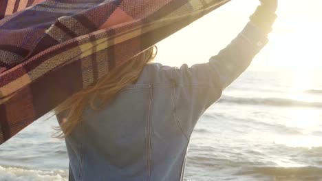 Young-beautiful-woman-raises-hands-up-with-plaid-on-the-shore-of-the-sea.-Happy-female-enjoying-the-sunset-on-the-beach