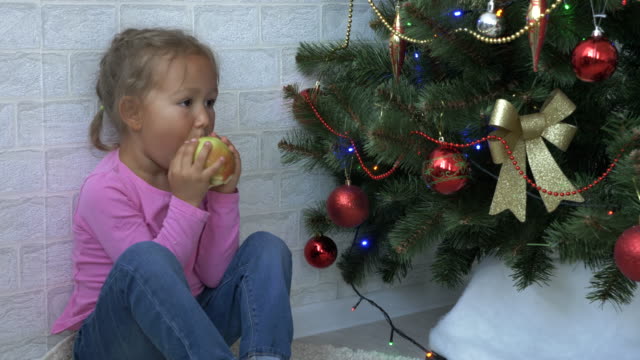 Niedliche-kleine-Mädchen-sitzen-auf-dem-Boden-und-Essen-Apfel-neben-einem-Weihnachtsbaum.