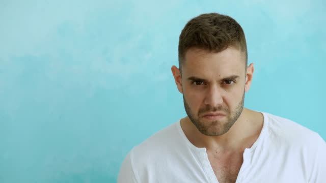 Portrait-of-angry-young-man-looking-into-camera-on-blue-background