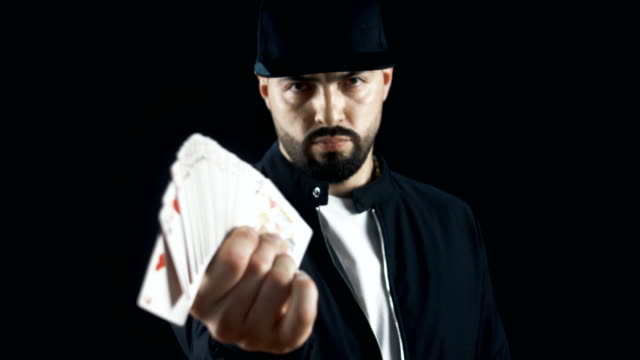 Professional-Street-Magician-in-a-Cap-Performing-Sleight-of-Hand-Card-Tricks,-Fan-Out-Cards,-Spinning-One-Card-on-a-Finger.-Background-is-Black.