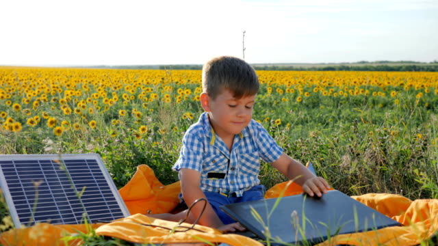 Kind-mit-Solarbatterie-lädt-Laptop-auf-Hintergrund-Feld-von-Sonnenblumen,-glückliches-Kind-schaut-Notebook-mit-solar-Ladegerät