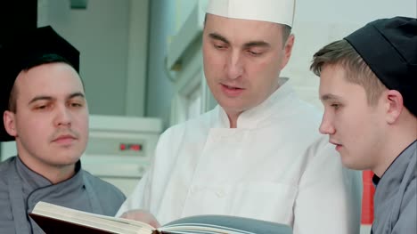 Jefe-de-cocina-sosteniendo-el-libro-de-recetas-y-hablar-de-algo-con-sus-aprendices