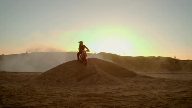 Long-Shot-Of-the-Extreme-Motocross-Rider-in-a-Cool-Protective-Helmet-Standing-on-the-Sand-Dune-in-the-Middle-of-Scenic-Quarry-with-Mist-and-Dust-Covering-Him.
