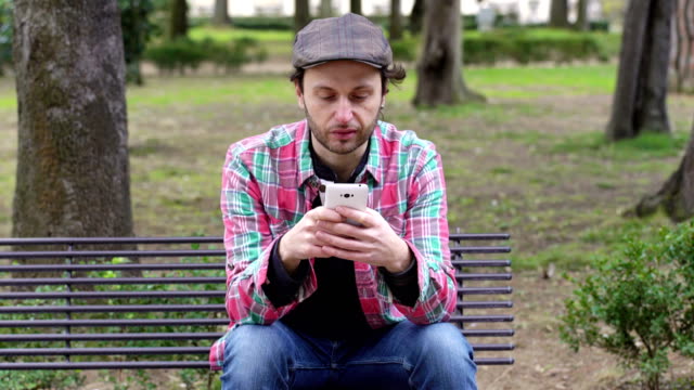 joven-sentado-en-un-banco-en-el-parque,-charla-alegremente-con-su-smartphone