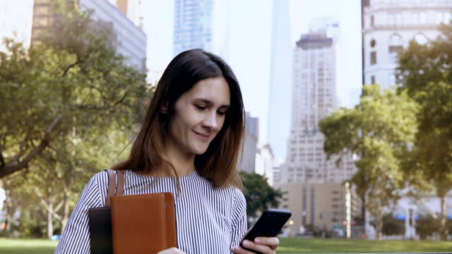 Young-happy-businesswoman-standing-in-financial-district-of-New-York,-USA-and-using-the-smartphone-as-mobile-office
