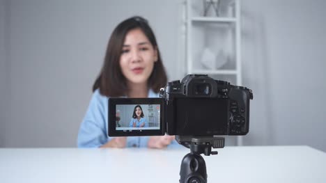 Feliz-sonriente-blogger-mujer-o-belleza-asiática-con-cepillo-y-cámara-de-grabación-de-video-y-agitando-la-mano-en-casa.-Concepto-de-belleza-videoblog-blog-personas.-Carro-de-tiro.