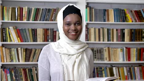 Young-african-muslim-girl-in-hijab-is-holding-book,-watching-at-camera,-religioun-concept,-booksheves-on-background
