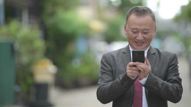 Reife-japanischer-Geschäftsmann-mit-Telefon-in-den-Straßen-im-freien