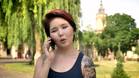 Retrato-de-jóvenes-mujeres-asiáticas-con-tatuada-mano-hablando-por-teléfono-y-de-pie-cerca-de-la-Universidad-en-el-parque,-sonriendo