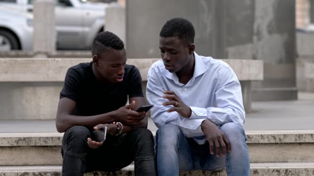 junge-schwarze-afrikanische-Freunde-sprechen-mit-Handys-in-Händen---outdoor