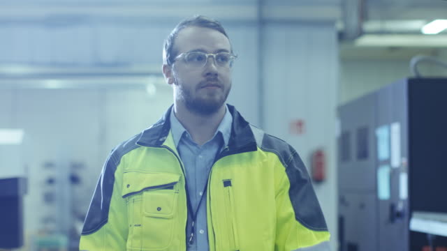 Industrial-Engineer-Wearing-Protective-Clothing-Puts-on-Hard-Hat-and-Walks-Through-Modern-Manufacturing-Facility-with-Automatic-Machinery-Working-in-Background.