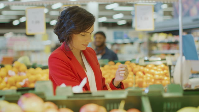 At-the-Supermarket:-Portrait-of-the-Beautiful-Smiling-Woman-Choosing-Products-In-the-Fresh-Produce-Aisle-and-Places-them-into-Shopping-Basket.-In-the-Background-Colorful-Fruits-and-Organic-Vegetables.-Slow-Motion.