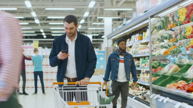 At-the-Supermarket:-Handsome-Man-with-Smartphone,-Pushes-Shopping-Cart,-Walks-Through-Fresh-Produce-Section-of-the-Store,-Chooses-Some-Products.-Other-Customers-Purchasing-Products.