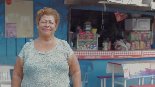 A-wide-portrait-of-an-older-hispanic-woman-looking-at-the-camera-standing-in-front-of-a-food-stand-in-Mexico
