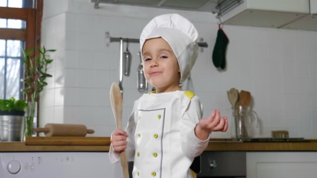 Ein-kleines-Mädchen-gekleidet-wie-ein-professioneller-Koch-in-der-Küche-mit-einem-Holzlöffel-als-Mikrofon-singt.
