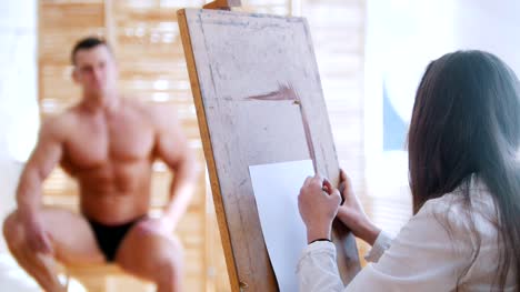 Young-woman-sits-behind-handsome-male-shirtless-model-and-drawing-him