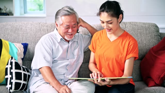 Daughter-reading-book-to-her-old-man-father-in-living-room.-Asian-senior-man-with-white-beard-and-asian-woman.-Senior-lifestyle-family-concept.