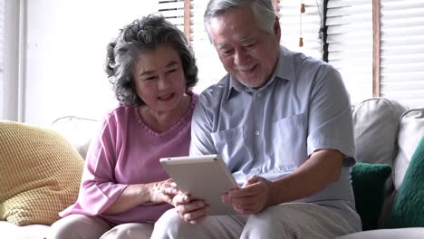 Senior-pareja-sujetando-el-tablet-y-sentados-en-el-sofá-juntos-en-la-sala-de-estar-en-casa.-Concepto-de-familia-feliz,-post-jubilación,-calidad-vida,-salud-mental-y-psicología-positiva.-resolución-de-4-k.