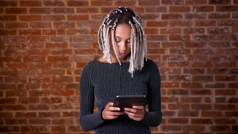 Young-African-girl-with-dreadlocks-using-a-tablet-computer,-typing.-Brick-wall-in-the-background.