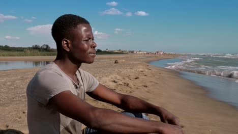 einsamer-junger-schwarzen-afrikanischen-Mann-sitzt-am-Strand,-das-Meer-betrachten