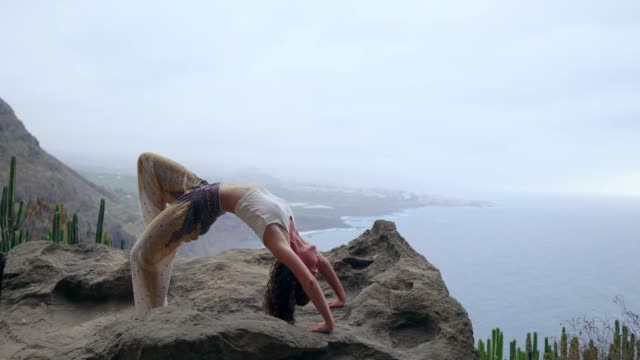 Mädchen-üben-von-Yoga-auf-den-Felsen-gegen-den-blauen-Himmel-und-das-azurblaue-Meer.-Frau-steht-auf-einem-Stein-in-einer-Brücke-Haltung.