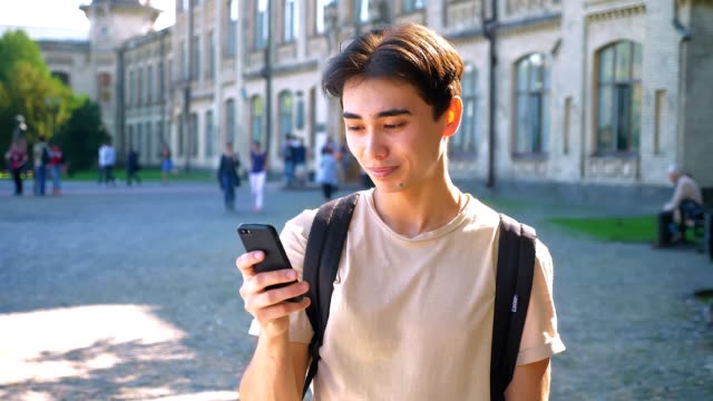 Asian-good-looking-male-is-scrolling-his-phone-and-glancing-at-camera-chill,-outdoor,-urban-mood