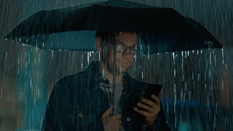 Young-Caucasian-Man-in-Glasses,-Wearing-a-Jeans-Coat-and-Square-Shirt-is-Using-a-Smartphone-Under-an-Umbrella.-It's-Dark-Outside-and-it-is-Raining.
