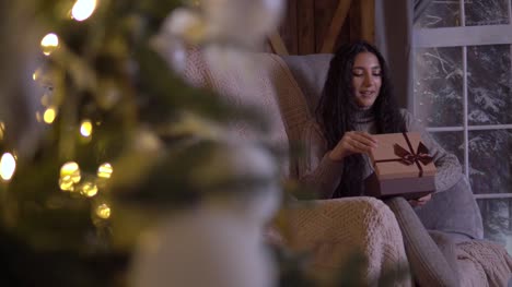 Das-Mädchen-öffnet-die-Box-mit-einem-Geschenk-und-freut-sich,-sitzt-auf-einem-Stuhl-in-der-Nähe-der-Weihnachtsbaum-am-Weihnachtstag