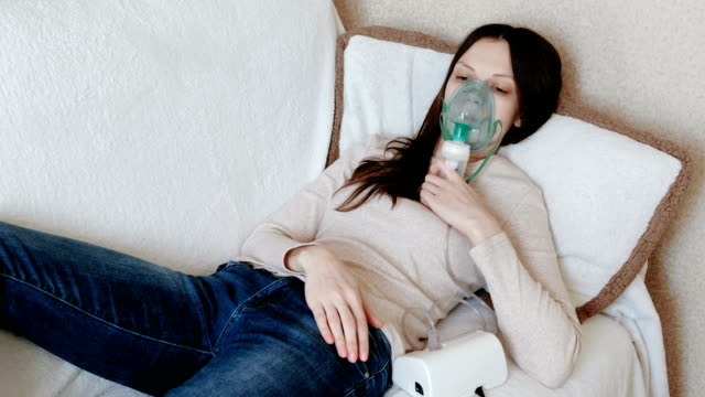 Utilice-el-nebulizador-y-el-inhalador-para-el-tratamiento.-Mujer-joven-inhalando-a-través-de-mascarilla-inhalador-tumbado-en-el-sofá.-Vista-frontal.