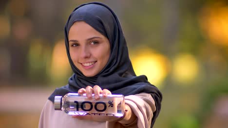 Primer-plano-retrato-de-muchacha-musulmana-en-hijab-mirando-a-cámara-con-sonrisa,-mostrando-la-botella-de-agua