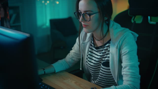 Excitado-y-bastante-Gamer-chica-en-gafas-es-jugar-videojuego-Online-en-su-ordenador-Personal.-Sala-y-PC-tienen-caliente-neón-Led-luces-de-colores.-Agradable-noche-en-la-casa.