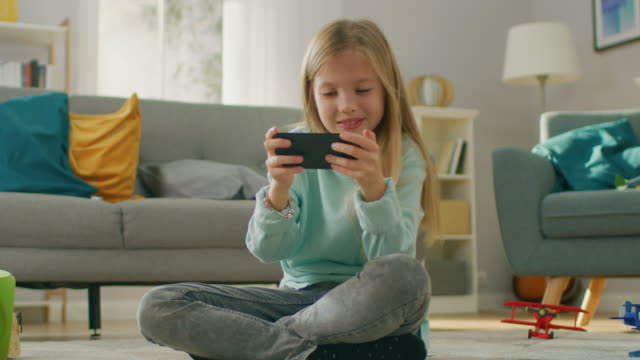 Intelligente-niedliche-Mädchen-sitzt-auf-einem-Teppich-zu-Hause-spielen-im-Videospiel-auf-seinem-Smartphone,-hält-und-nutzt-Handy-im-horizontalen-Landscape-Modus.-Kind-hat-Spaß-im-sonnigen-Wohnzimmer-Videospiel-zu-spielen.-In-Zeitlupe.