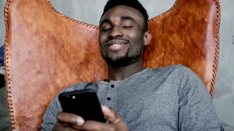 Afrikanische-männlichen-im-Stuhl-sitzen-und-denken,-hält-Smartphone.-Mann-merkt-sich-etwas-gutes-und-beginnt,-Eingabe-von-Nachrichten