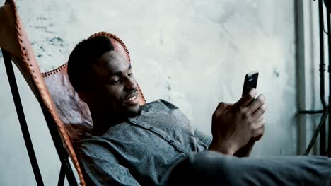 Portrait-des-afrikanischen-Mann-sitzt-im-Stuhl-mit-einem-Smartphone.-Hübscher-Mann-lächelt-und-schaut-Fotos-auf-seinem-Handy