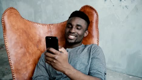 Portrait-of-African-man-sitting-in-the-chair-and-using-the-Smartphone.-Male-is-surfing-the-internet,-smiling