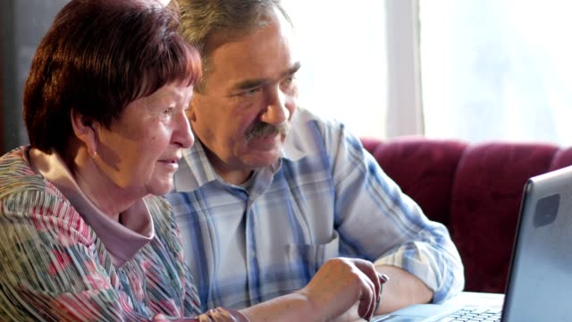 An-elderly-couple-is-sitting-at-home-at-the-laptop.-A-woman-reads-news,-a-man-with-a-mustache-sits-next-to-him-and-talks